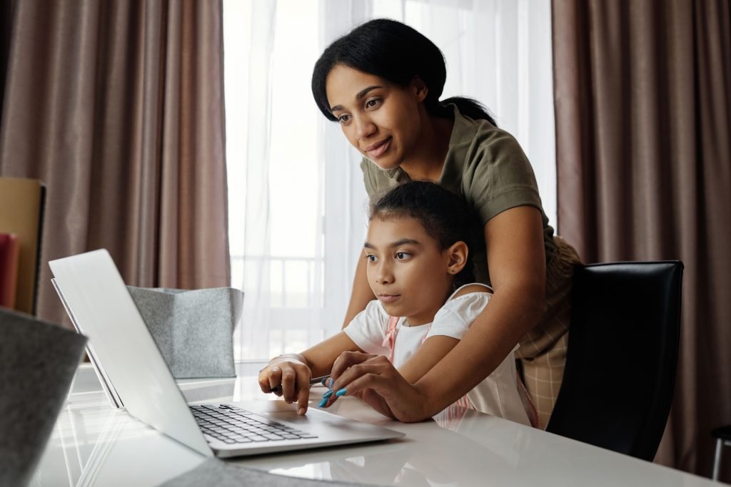 A parent standing beside a child as they work together at a laptop at home.